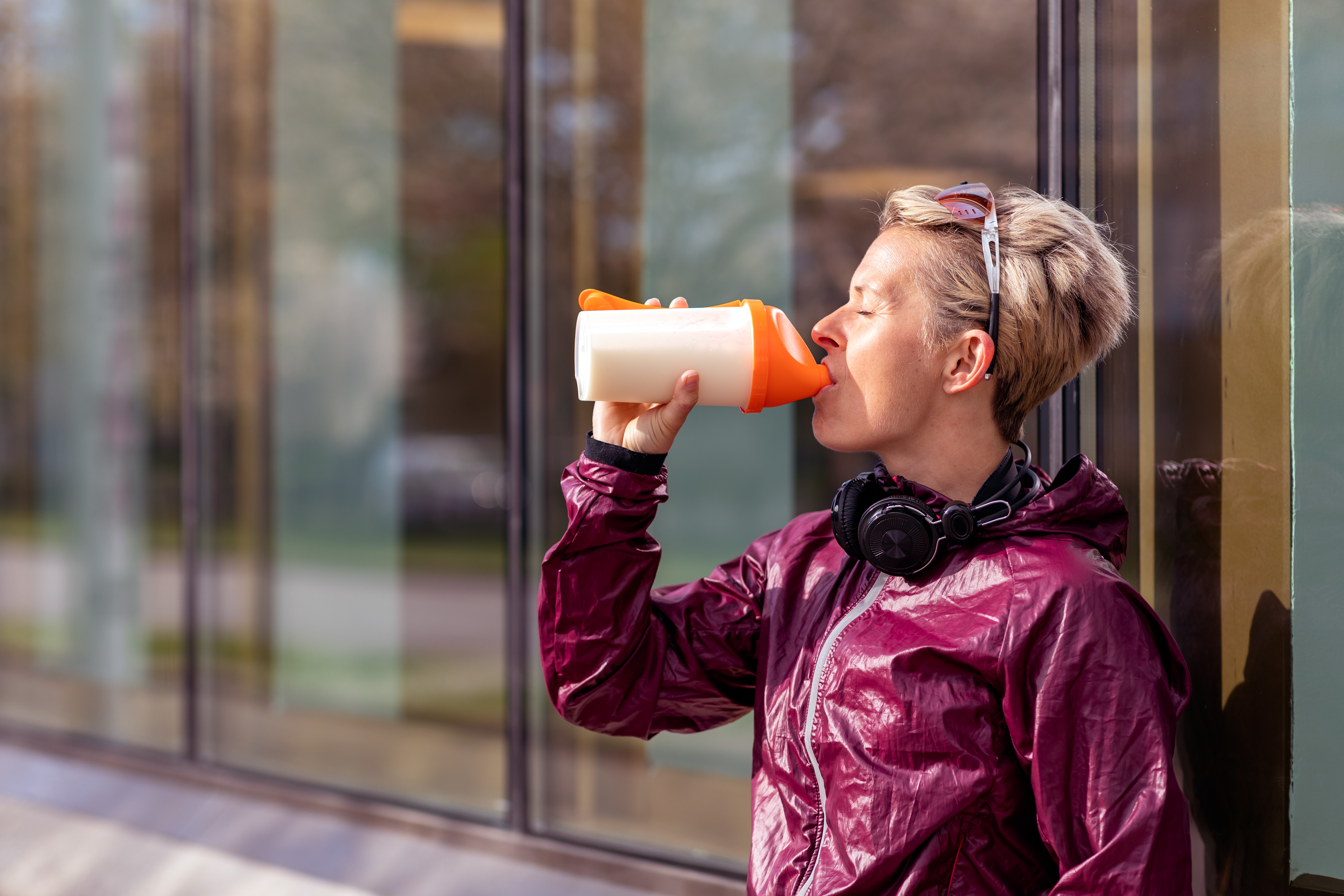 A woman in sports clothing drinking a protein shake. She is wearing a pair of headphones around her neck and a pair of sunglasses on her head. (Foto: Getty Images)