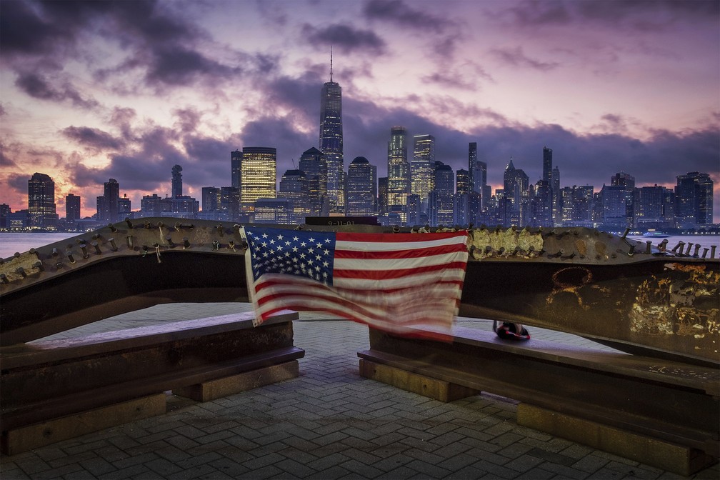 Uma bandeira dos EUA é vista sobre ferragem retorcida restante do ataque terrorista às Torres Gêmeas em 2001, em Jersey City, com os prédios de Nova York ao fundo, no dia que marca os 18 anos dos atentados de 11 de Setembro — Foto: J. David Ake/AP