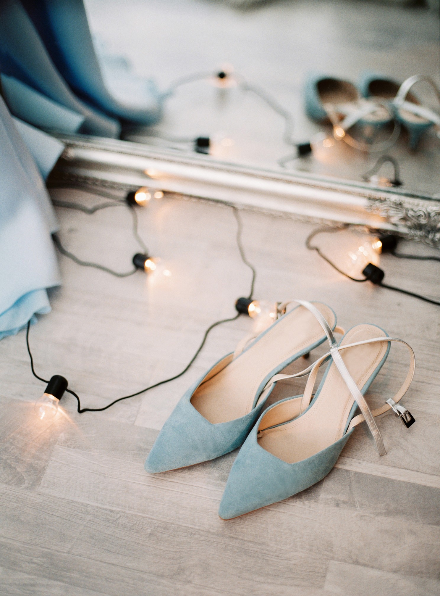Blue suede high heel summer female shoes and lamps garland on a floor, with mirror background, contemporary fashion. Top view, flat lay, closeup. (Foto: Getty Images/iStockphoto)