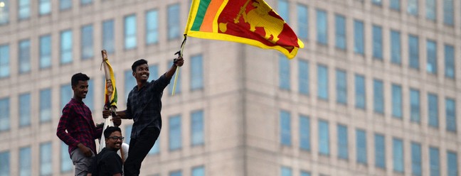 Homem agita a bandeira nacional do Sri Lanka depois de subir em uma torre perto da secretaria presidencial, na capital Colombo, em protesto contra o governo. O presidente Gotabaya Rajapaksa fugiu, deixando para trás milhões de rúpias em dinheiro que foram entregues à polícia — Foto: ARUN SANKAR / AFP