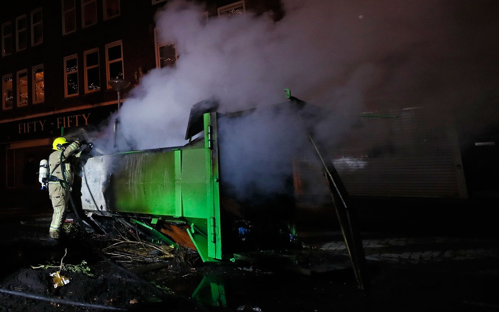 Bombeiro extingue chamas de conteiner de lixo incendiado durante protesto contra toque de recolher em Roterdã, na Holanda, na segunda-feira (25) — Foto: AP Photo/Peter Dejong