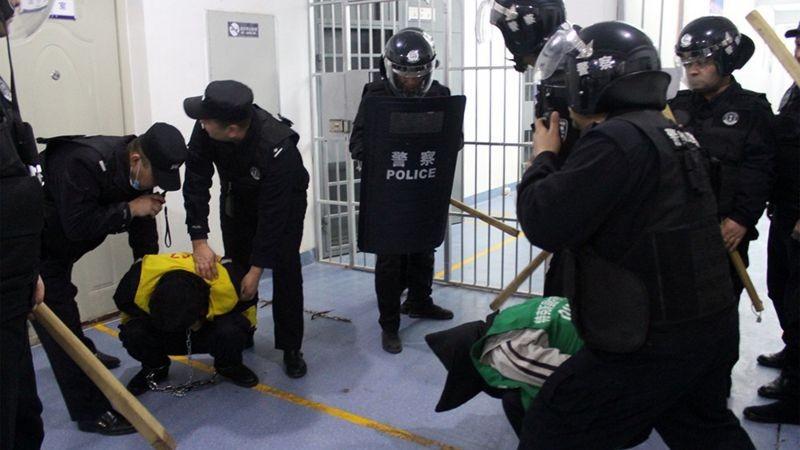 Um homem detido aparece agachado, de cabeça baixa, rodeado por policiais com cacetetes (Foto: BBC News)