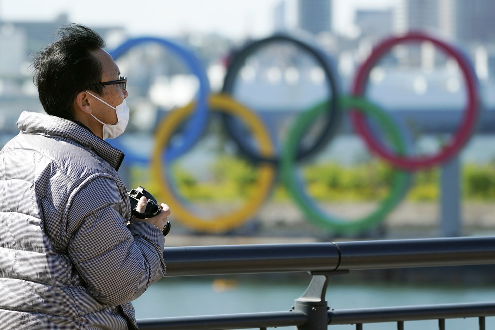 Homem usando máscara perto dos anéis olímpicos em Tóquio, no distrito de Odaiba, após o adiamento da Olimpíada — Foto: Eugene Hoshiko/AP