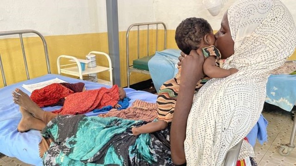 Hawa segura sua filha Ubah ao lado da cama onde seu filho Abdiwali está deitado — Foto: BBC/ ED HABERSHON