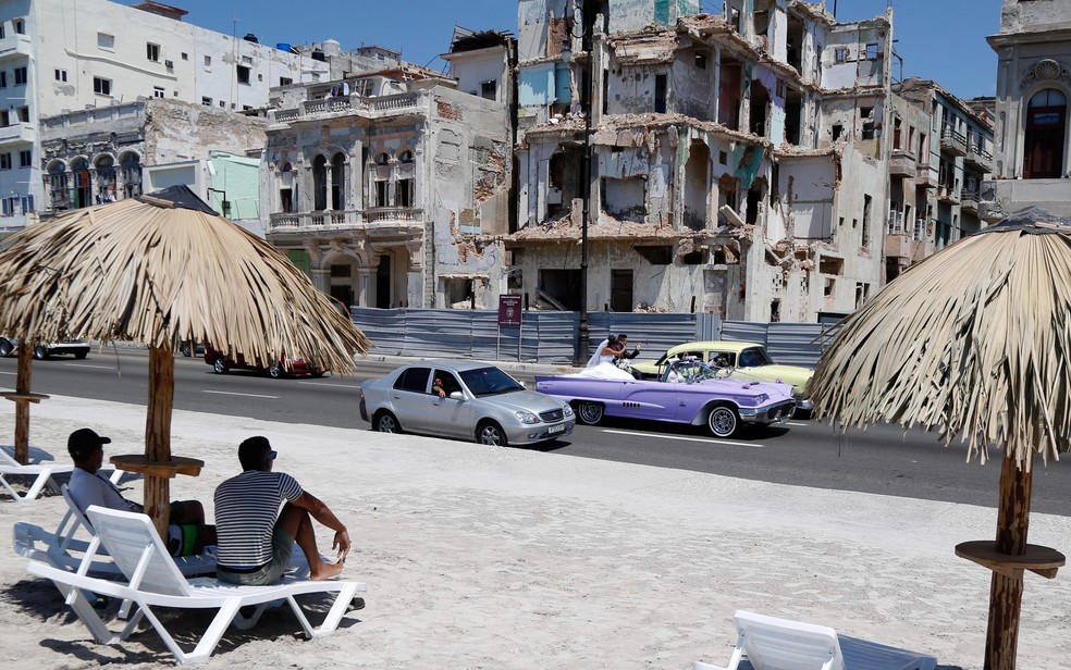 Pessoas se sentam em uma praia artificial enquanto um casal de noivos passa em um carro conversível em Havana, Cuba.  — Foto: Desmond Boylan/AP
