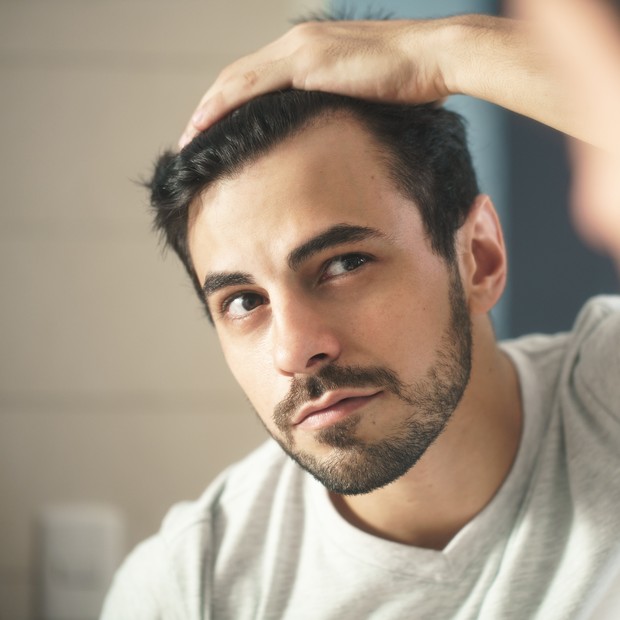 Latino person with beard grooming in bathroom at home. White metrosexual man worried for hair loss and looking at mirror his receding hairline. (Foto: Getty Images/iStockphoto)
