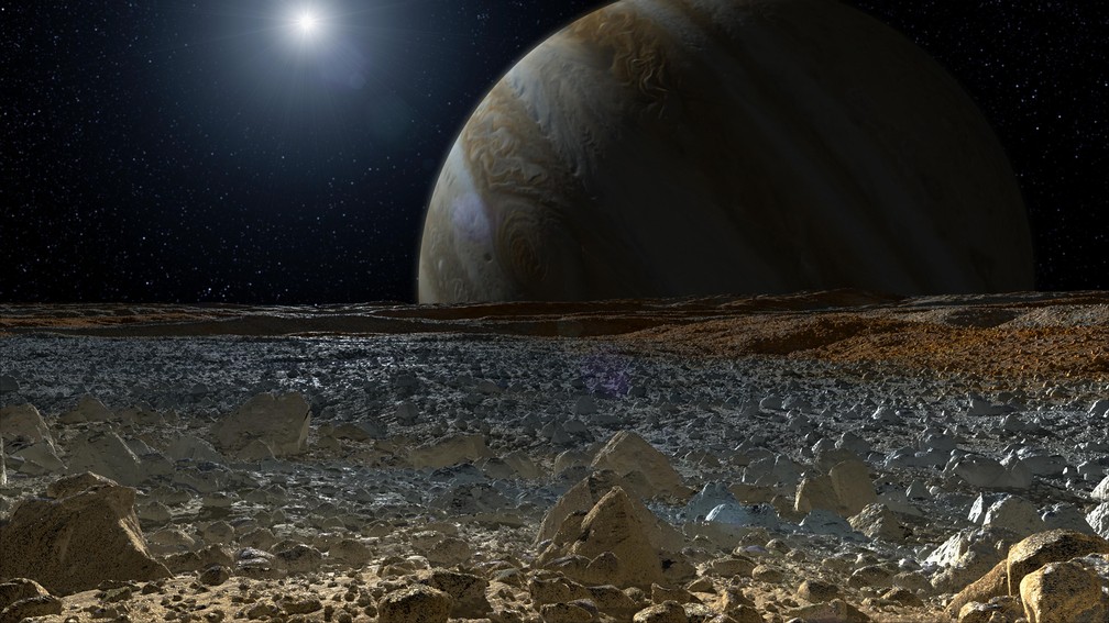 Representação artística da superfície de Europa, uma das luas de Júpiter. Ao fundo, o gigante gasoso está em destaque. — Foto: NASA/JPL-Caltech