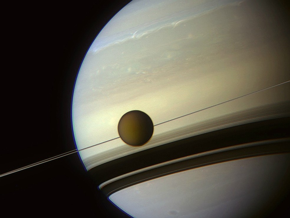  Maior lua de Saturno, Titã orbita o planeta em imagem captada pela sonda Cassini, da Nasa. Os anéis de Saturno são planos e vistos como uma linha fina, parecendo 'espetar' Titã.  — Foto: NASA/JPL-Caltech/Space Science Institute/J. Major 