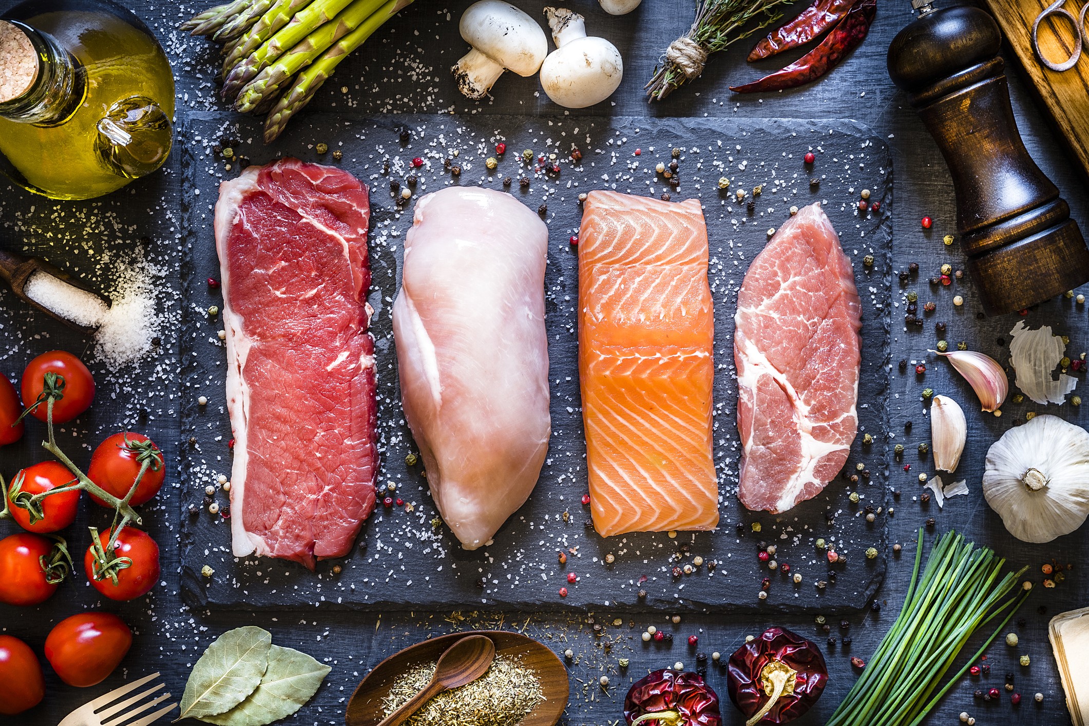 Top view of four different types of animal protein like a raw beef steak, a raw chicken breast, a raw salmon fillet and a raw pork steak on a stone tray. Stone tray is at the center of the image and is surrounded by condiments, spices and vegetables. Low  (Foto: Getty Images)