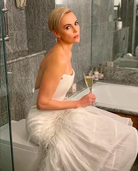 A atriz Charlize Theron fazendo graça com seu vestido de gala e uma taça de champagne, sentada em uma privada de um evento em Nova York (Foto: Instagram)