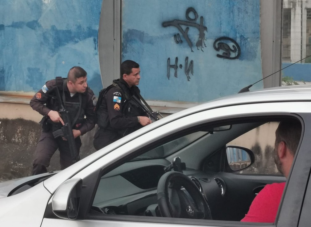 Dupla de policiais, com fuzil na mão, tenta olhar por trás do muro que separa a Linha Vermelha da Maré — Foto: Marcos Serra Lima/g1