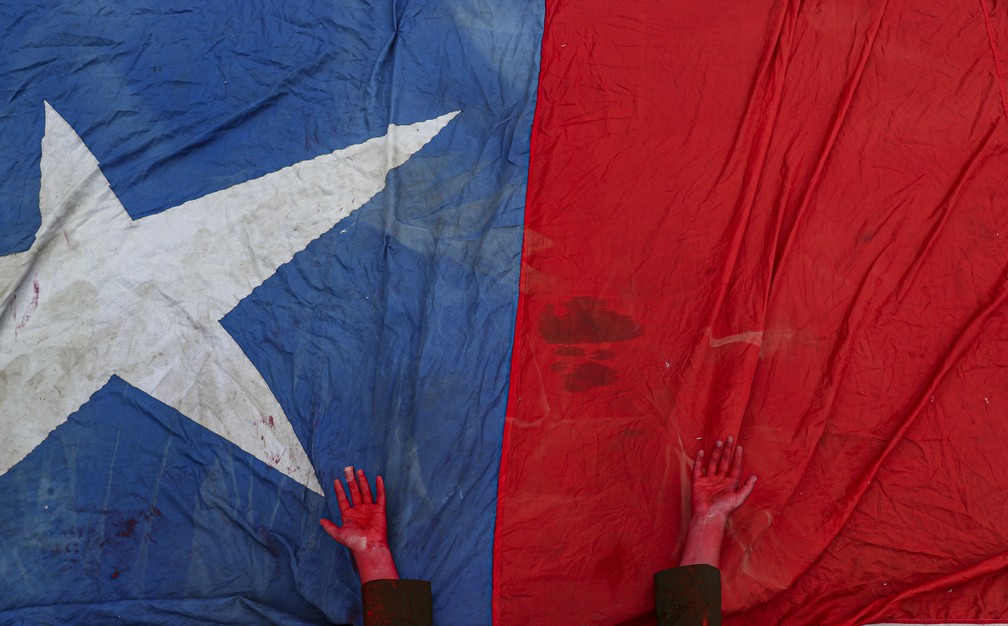 Manifestante com mão pintada de vermelho para simbolizar mortes nos protestos do Chile em bandeira durante protesto em Santiago neste domingo (27) — Foto: Esteban Felix/AP Photo