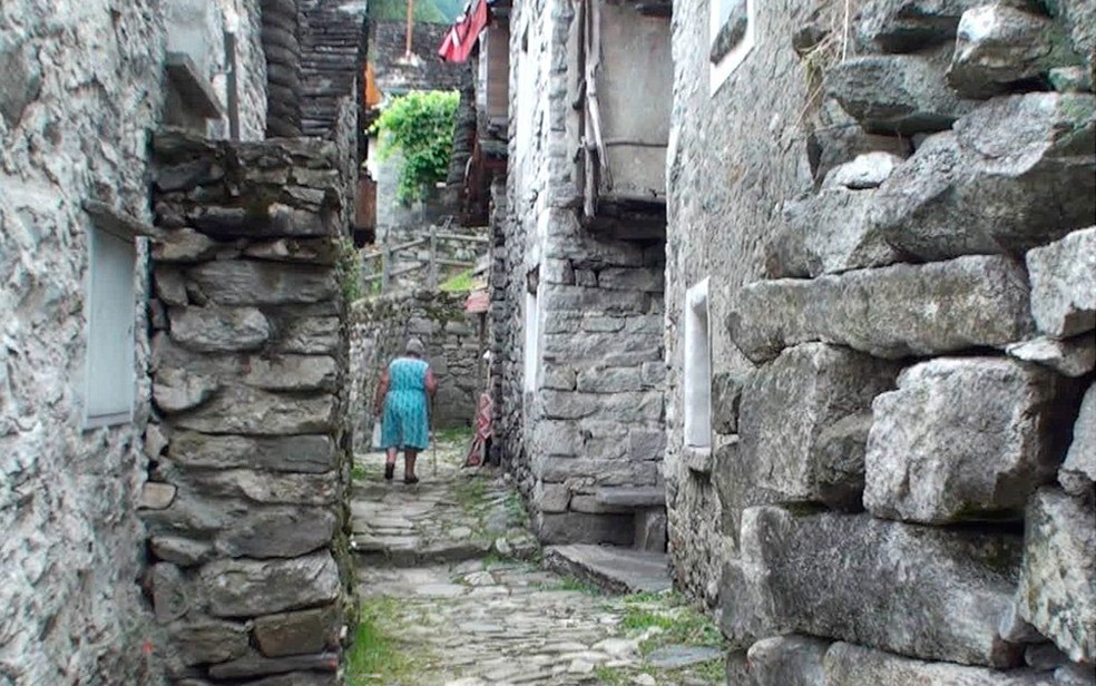 Senhora caminha por uma rua estreita de Corippo, ladeada por construções de pedra (Foto: BBC)