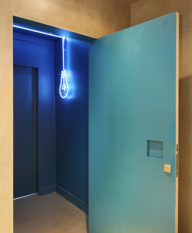HALL | Na entrada do elevador, o destaque fica por conta da luminária com filamento de neon, da Neon 3 Estações, desenhada pelo escritório, em contraste com a pintura azul de todas as paredes e teto, formando um grande bloco (Foto: Mariana Orsi / Divulgação)