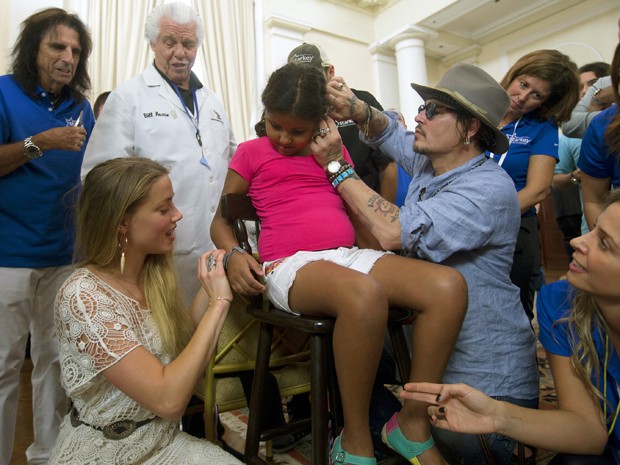 O ator Johnny Depp coloca um aparelho auditivo no ouvido de uma criança enquanto sua mulher, a atriz Amber Heard, dá uma pulseira à criança (Foto: AP Photo/Silvia Izquierdo)