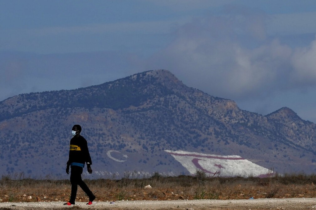 Migrante anda em frente a montanha com a bandeira do Chipre do Norte, território de maioria turca no Chipre e que gera disputa política, em foto de 25 de novembro — Foto: Petros Karadjias/AP Photo