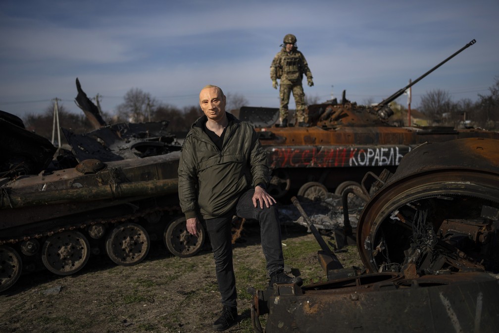 7 de abril - Um civil usa uma máscara de Vladimir Putin, enquanto um soldado ucraniano fica em cima de um tanque russo destruído em Bucha, Ucrânia — Foto: Rodrigo Abd/AP