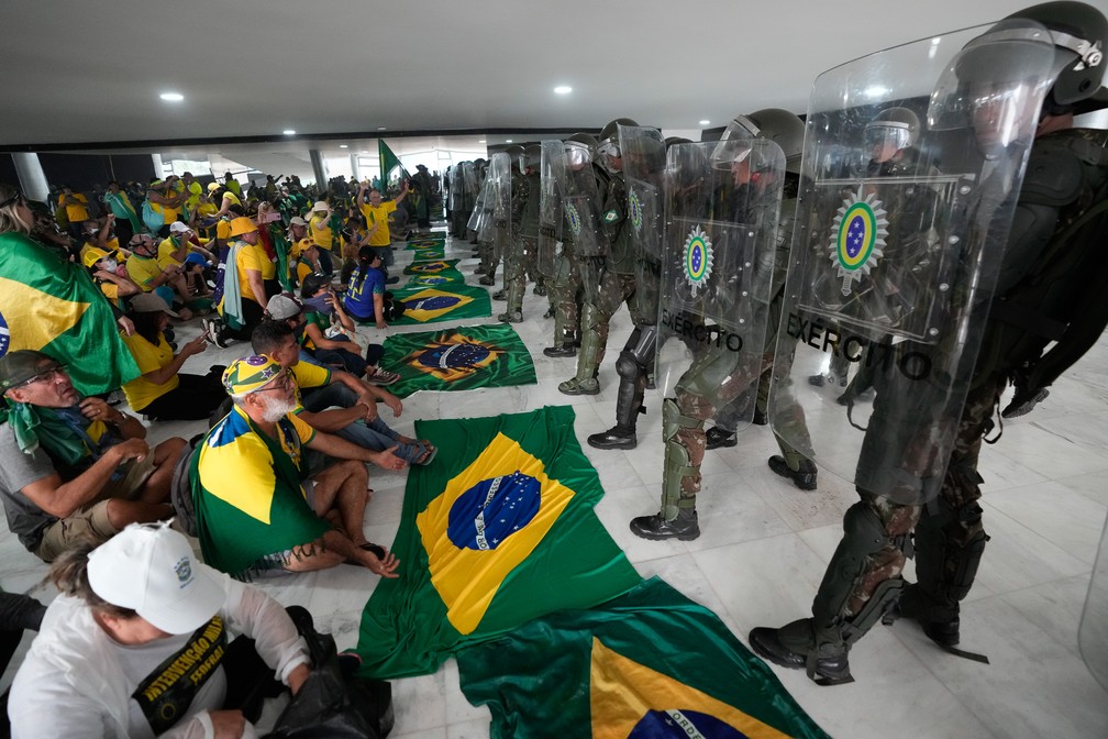 Invasores do Palácio do Planalto se sentam com bandeiras do Brasil diante de militares — Foto: Eraldo Peres/AP