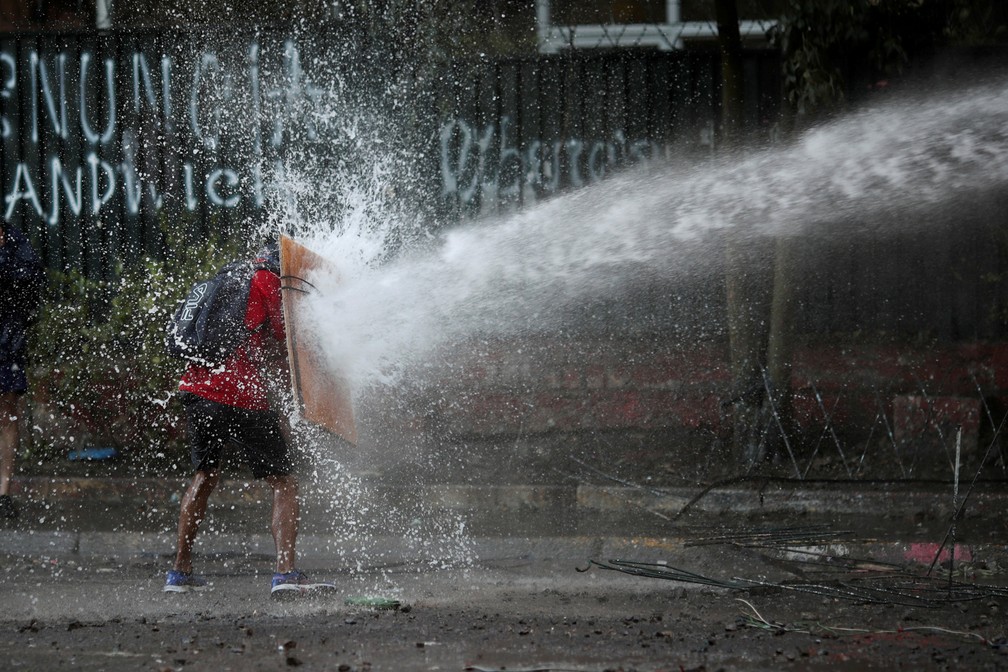 12 de dezembro - Um manifestante segura um pedaço de madeira como escudo improvisado para se proteger de um canhão d'água disparado pela tropa de choque da polícia durante protesto contra o governo em Santiago, no Chile — Foto: Ricardo Moraes/Reuters