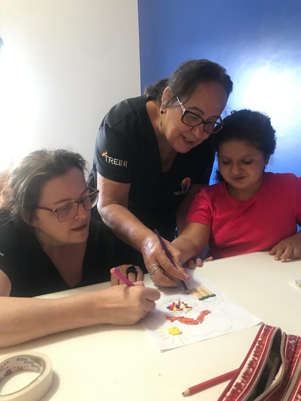 Foto: Adriana (psicóloga, sentada) e Manoela (psicopedagoga, em pé) juntas com a pequena Letícia, de 10 anos, aplicando a metodologia MIG para trabalho de foco e concentração — Foto: Cedida