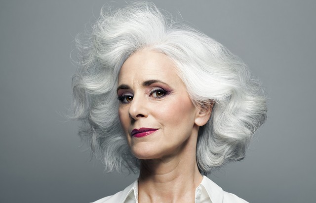 Mature woman with medium length, big, wavy, silvery, grey hair in front of grey background wearing make up red lip stick, portrait. (Foto: Getty Images)