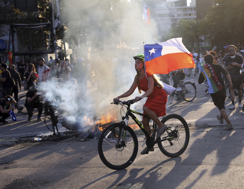 Mulher pedala com bandeira do Chile em frente a barricada em Santiago, onde ocorrem protestos neste sábado (26) — Foto: Rodrigo Abd/AP Photo