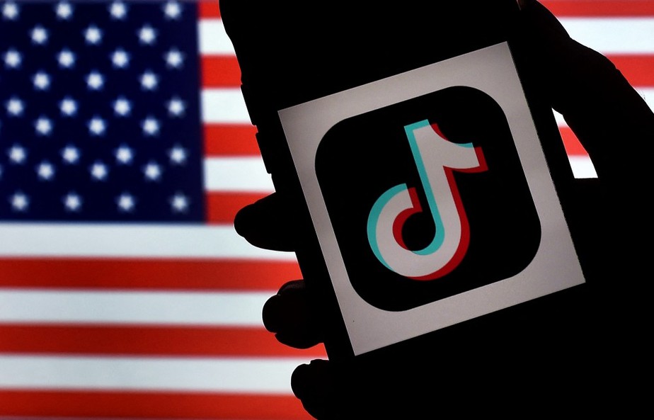 Foto ilustrativa mostra logo do TikTok em telefone na frente de painel com bandeira americana
