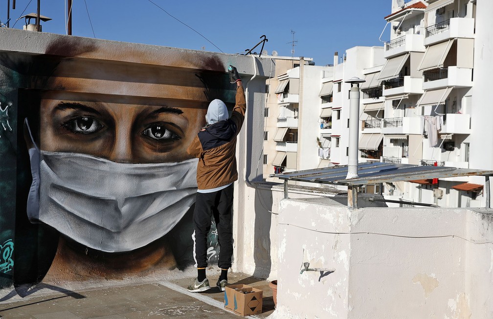 Um jovem grego de 16 anos de idade dá retoques no grafite de uma mulher usando uma máscara, inspirado na pandemia do coronavírus, no telhado do prédio onde ele mora em Atenas — Foto: Thanassis Stavrakis/AP