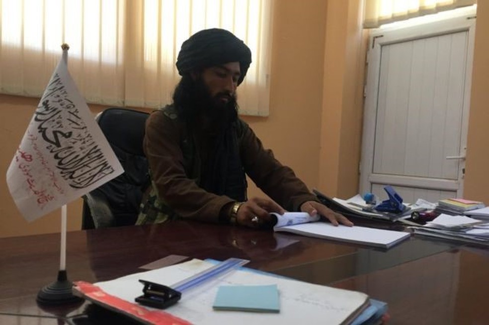 Prefeito do Talibã em Bactro em sua mesa de madeira, com uma pequena bandeira branca do "Emirado Islâmico do Afeganistão" — Foto: BBC