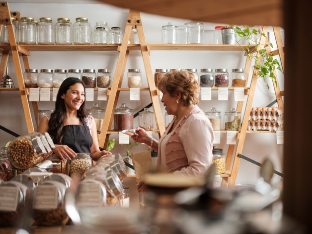 Happy hispanic employee and senior female customer standing next to the shelves of a sustainable shop and smiling at each other. (Foto: Getty Images)