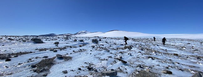Rochas espalhadas por um campo de gelo, com os cientistas procurando por meteoritos ao fundo — Foto: Maria Valdés