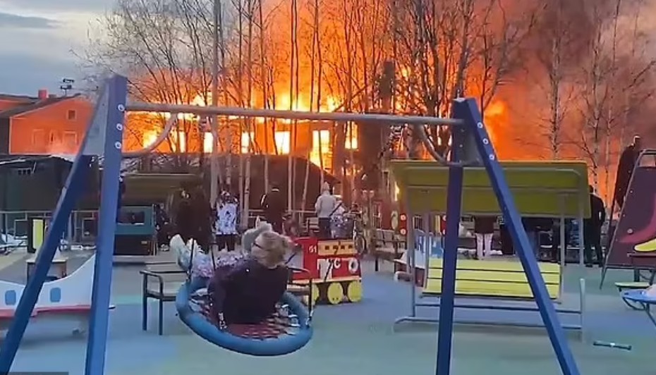 Mulher se embala com criança em playground enquanto prédio pega fogo ao fundo (Foto: Reprodução/Daily Mail)
