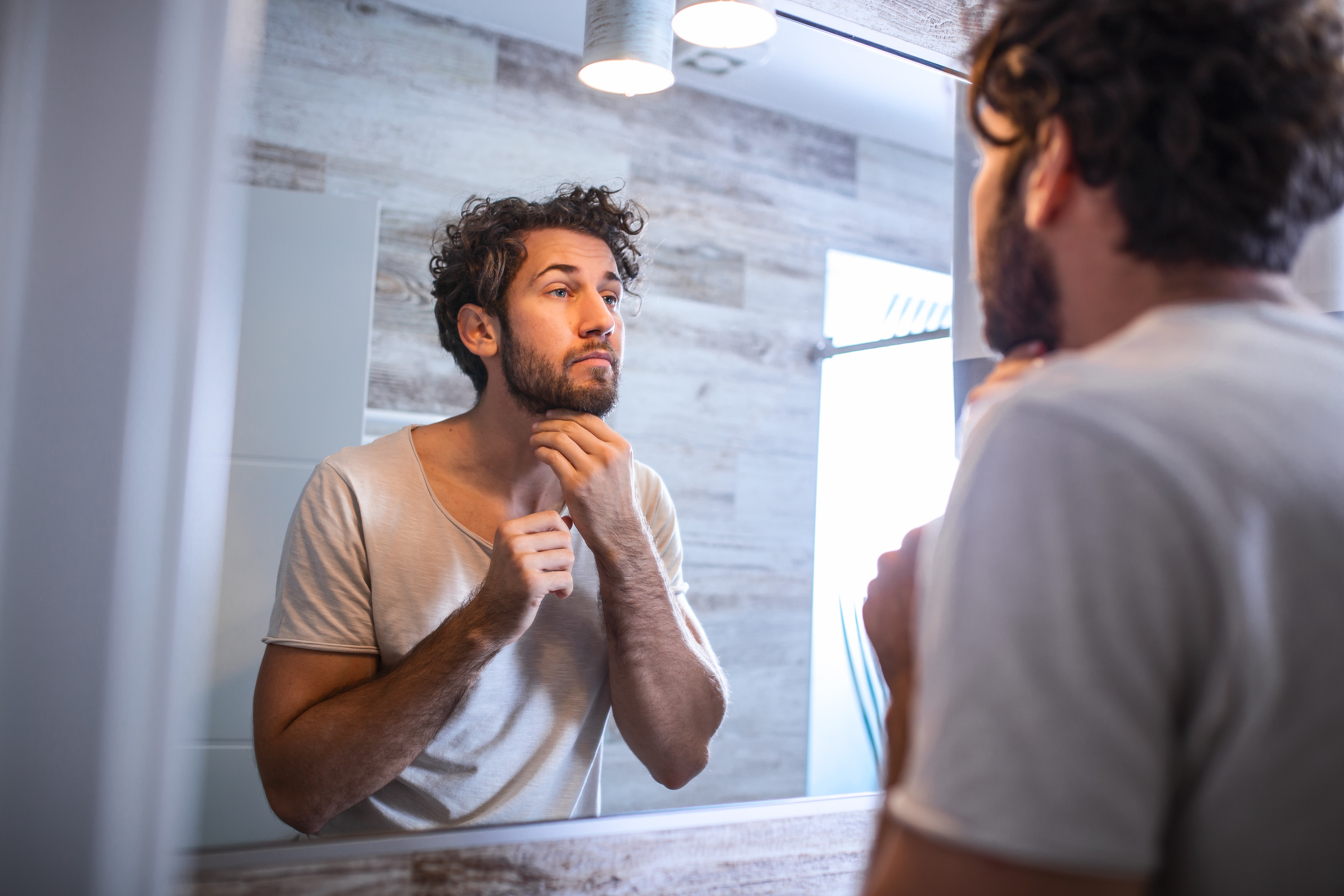 Reflection of handsome man with beard looking at mirror and touching face in bathroom grooming. Man putting skincare facial treatment cream on face. (Foto: Getty Images/iStockphoto)