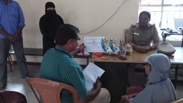 A menina juntamente com o pai prestando esclarecimentos à polícia. Detalhe para os troféus na mesa da autoridade (Foto: Reprodução BBC Tamil)