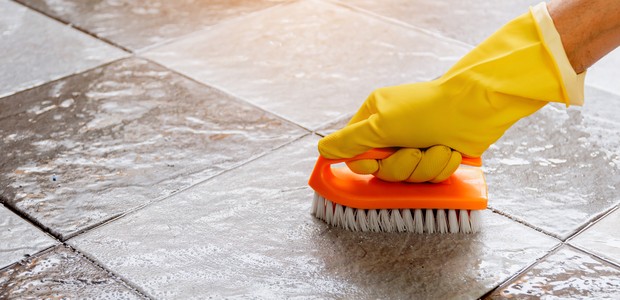 Hands wearing yellow rubber gloves are using a plastic floor scrubber to scrub the tile floor with a floor cleaner. (Foto: Getty Images/iStockphoto)